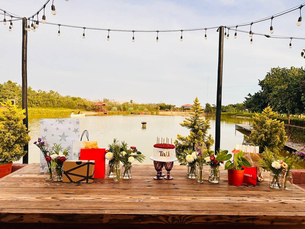 Mesa con flores en frascos, regalos y una torta frente a una laguna, bajo una guirnalda de focos