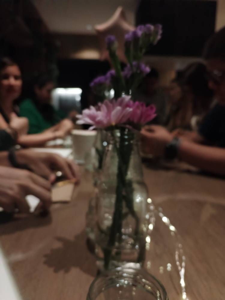 Flores en un frasco sobre una mesa de noche, con mujeres conversando detrás, fuera de foco