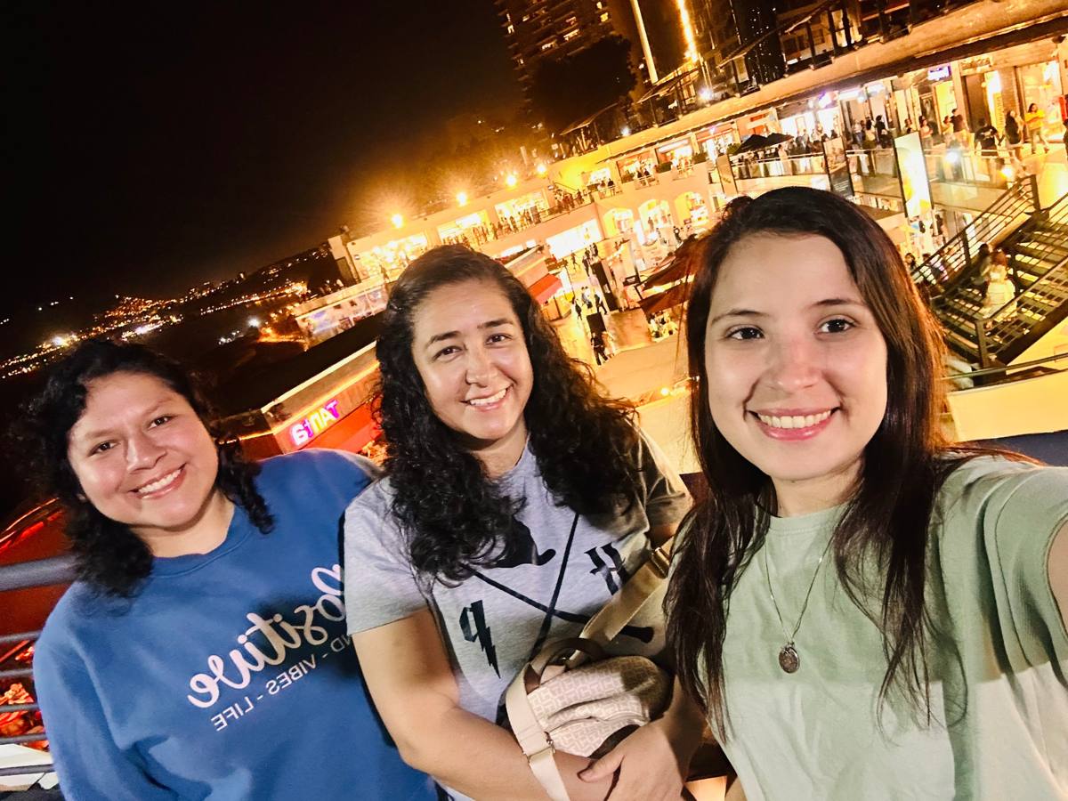 Tres mujeres sonríen en una selfie de noche, con luces cálidas de fondo